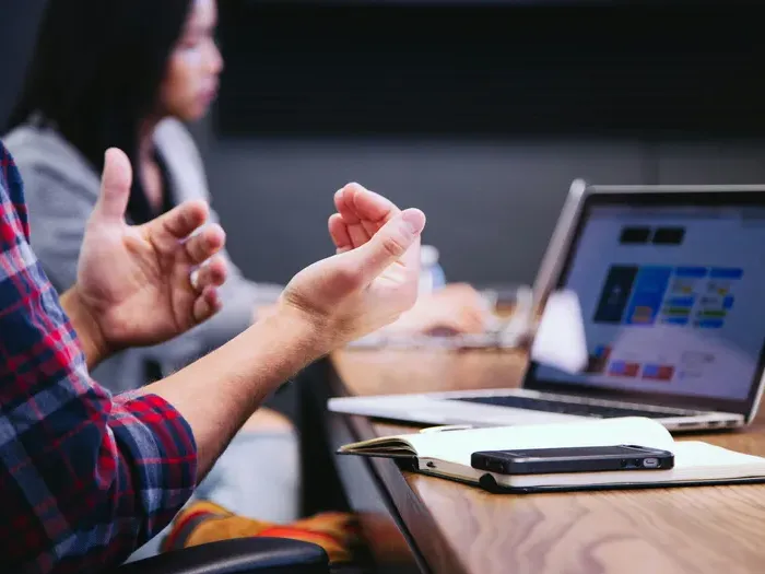 Two people in conversation with hands gesturing over desk with laptop and notebooks visible